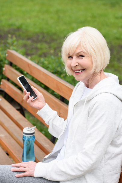 elderly cheerful woman using smartphone while sitting on bench with sport bottle of water - Foto, Imagem