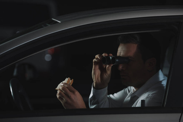 чоловічий приватний детектив робить спостереження за біноклями і їсть бутерброд в машині
 - Фото, зображення