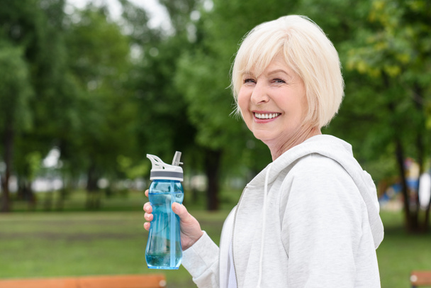mulher sênior segurando garrafa esporte e sorrindo para a câmera
 - Foto, Imagem