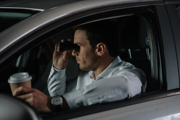 visão lateral de agente masculino disfarçado fazendo vigilância por binóculos e beber café no carro
 - Foto, Imagem