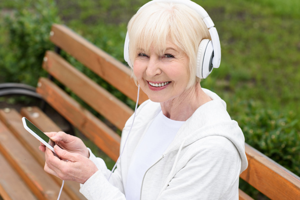 χαρούμενη ηλικιωμένη γυναίκα που ακούει μουσική με ακουστικά και smartphone - Φωτογραφία, εικόνα
