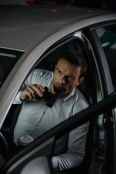 vista de ángulo alto de detective privado masculino con taza de café haciendo vigilancia por prismáticos desde el coche
 - Foto, Imagen