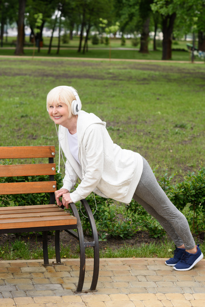 senior sportswoman in headphones doing push up near bench in park  - Photo, Image