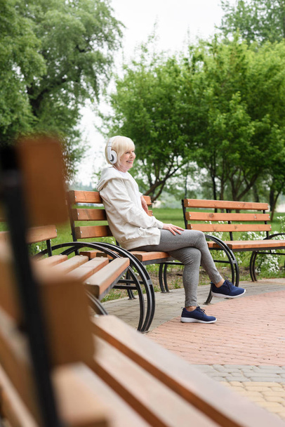 Senior kobieta słuchania muzyki w słuchawkach na ławce w parku - Zdjęcie, obraz