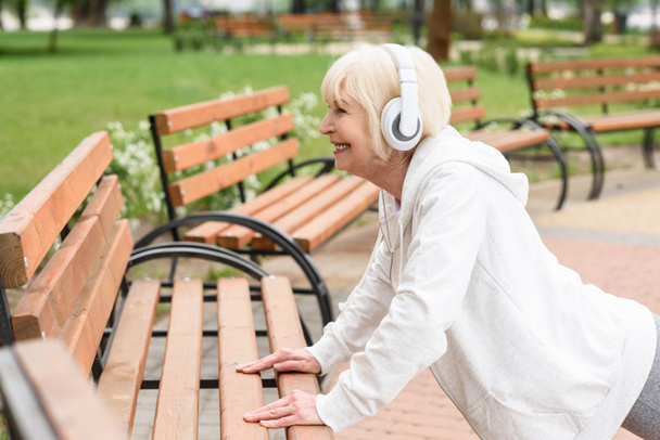 senior sportive woman in headphones training near benches in park - Photo, Image