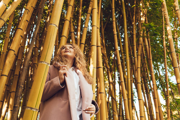 stylish young woman in pink coat walking in park and posing next to tall bamboo trees - Фото, изображение