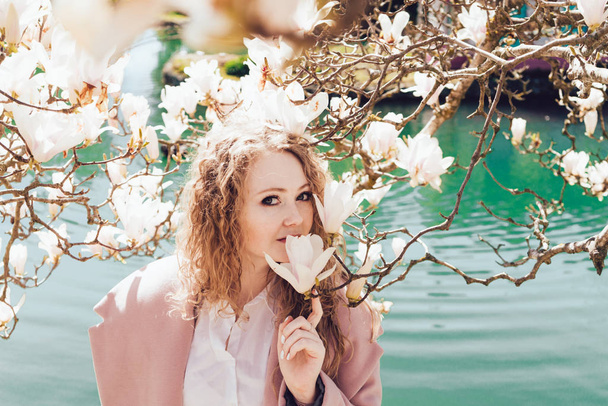 curly elegant girl enjoys the scent of a blooming magnolia in a park next to a pond, poses - Fotó, kép