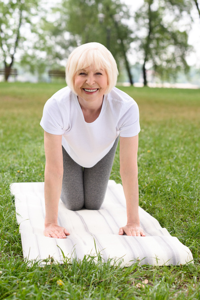 smiling elderly woman practicing yoga on mat on green lawn - Foto, afbeelding