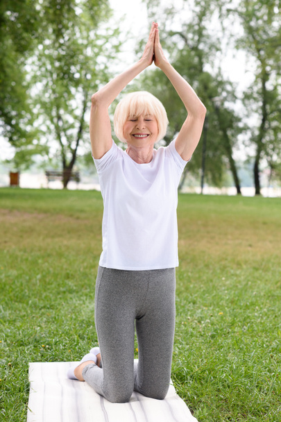 senior woman with namaste gesture practicing yoga on mat in park - Zdjęcie, obraz