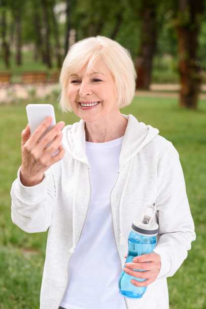 mujer mayor con botella de agua deportiva usando teléfono inteligente
 - Foto, Imagen