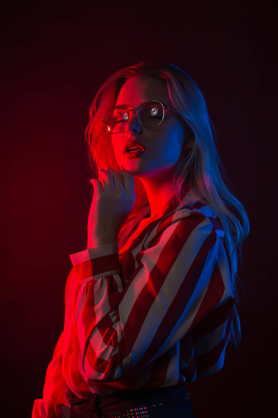Adorable young model in glasses wearing striped blouse, posing with red and blue light - Photo, Image