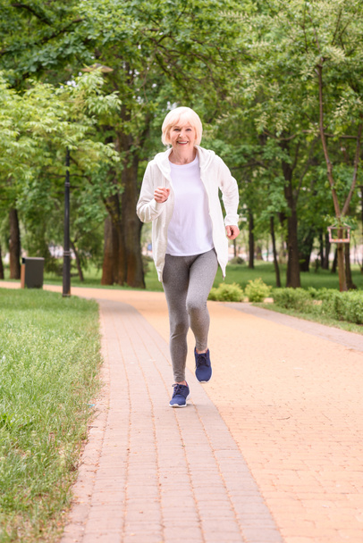 улыбающаяся старшая спортсменка, бегущая в парке
 - Фото, изображение