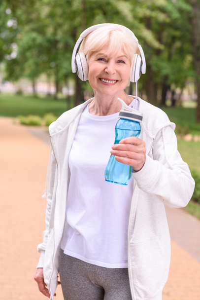 χαρούμενα αθλήτρια με μπουκάλι νερό και ακουστικά στέκεται στο πάρκο   - Φωτογραφία, εικόνα