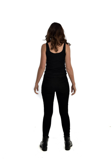 Ganzkörperporträt eines brünetten Mädchens mit schwarzem Singlet, Jeans und Stiefeln. Stehende Pose, mit dem Gesicht zur Kamera. isoliert auf weißem Studiohintergrund. - Foto, Bild