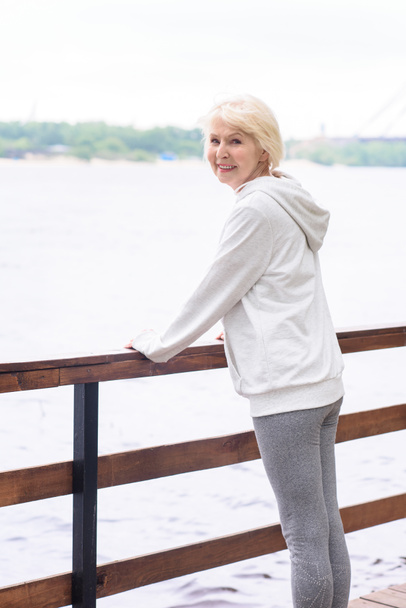 Glückliche Seniorin steht am hölzernen Geländer am Fluss  - Foto, Bild