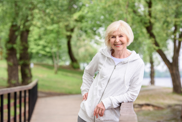 Lächelnde Seniorin steht neben Geländer im Park - Foto, Bild