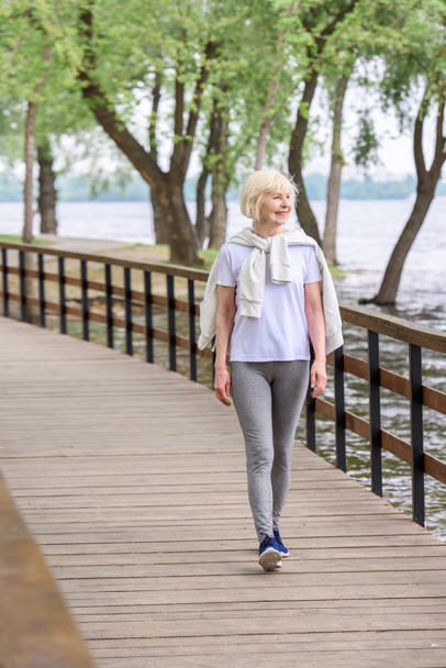 smiling senior woman walking on wooden path in park - Photo, Image