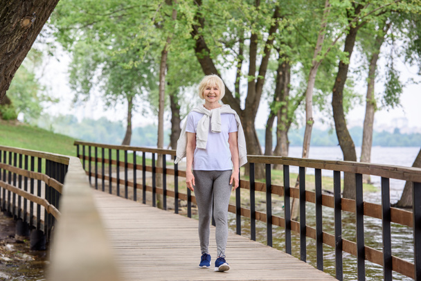 senior woman in sportswear walking on wooden path in park - Zdjęcie, obraz