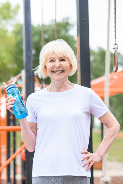 улыбающаяся старшая спортсменка с водой в спортивной бутылке, стоящая на спортивной площадке
  - Фото, изображение