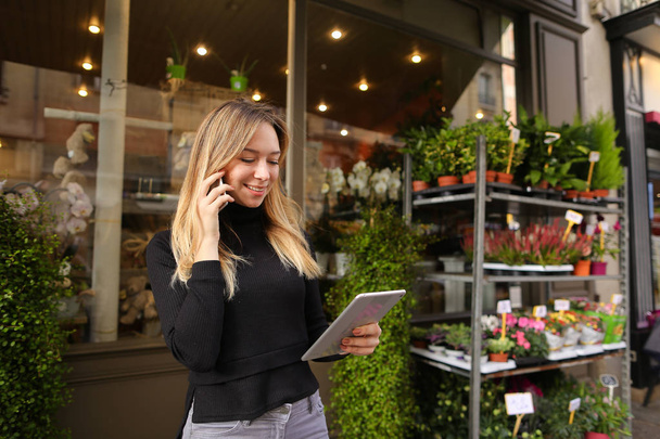 Vrouwelijke Amerikaanse manager van bloemenwinkel permanent met tablet en praten door smartphone. - Foto, afbeelding
