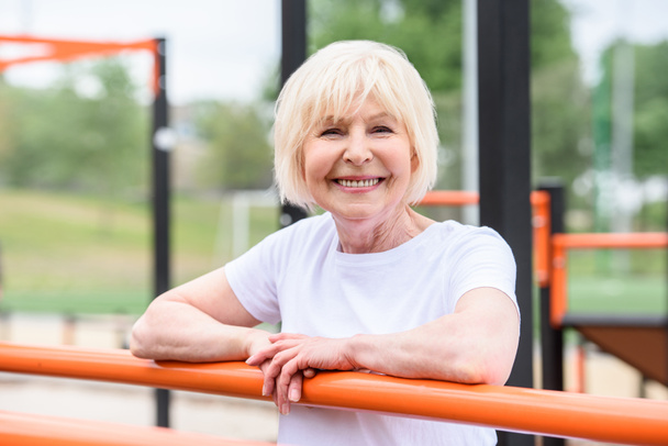 usměvavá starší žena stojící na sportoviště a při pohledu na fotoaparát - Fotografie, Obrázek