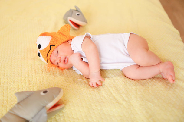 Маленький милый малыш лежит на желтом фоне с игрушками и в оранжевой кепке
. - Фото, изображение