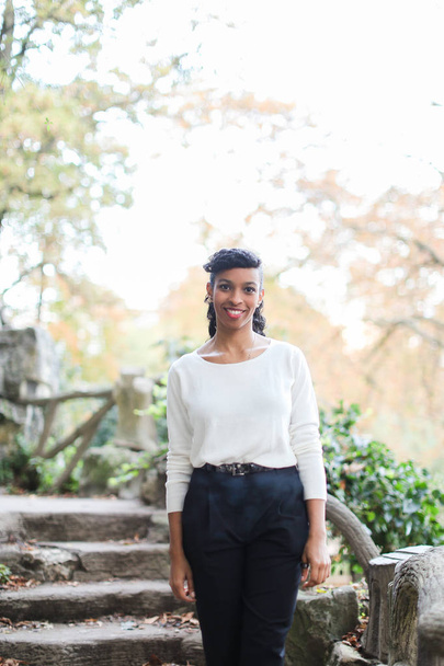 Sorridente ragazza nera che indossa camicetta bianca in piedi vicino a scale di pietra nel parco
. - Foto, immagini