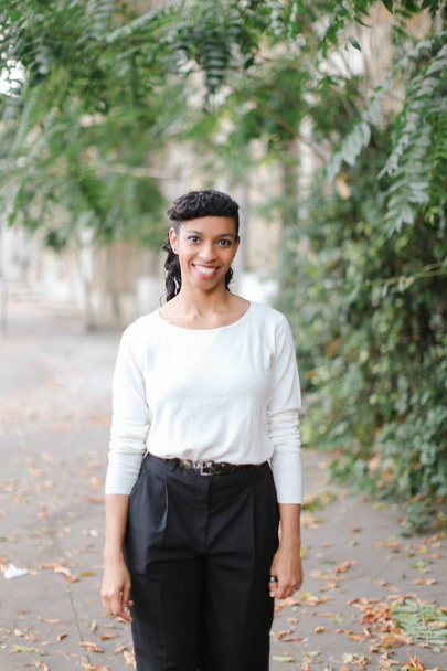Чорношкіра молода жінка, що йде біля будівлі та зелених дерев, носить білу блузку та чорні штани
. - Фото, зображення