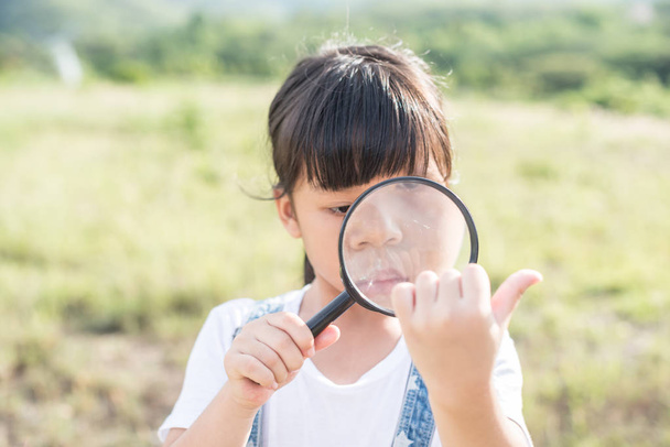 Little girl is playing on green meadow and examining field flowers using magnifying glass - Photo, Image