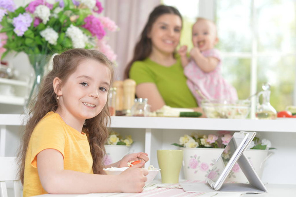 Petite fille mignonne mangeant de la salade fraîche à la table de cuisine avec tablette dessus
 - Photo, image