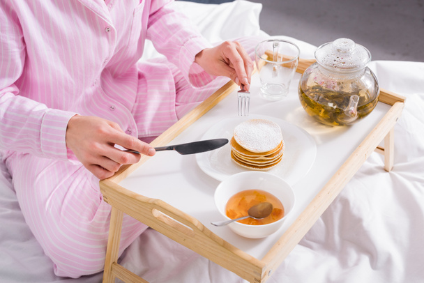 Teilbild einer Frau im Pyjama, die zu Hause Pfannkuchen mit Marmelade zum Frühstück im Bett isst - Foto, Bild