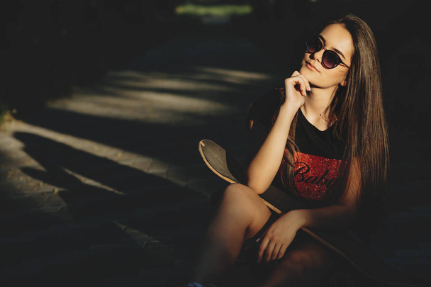 Εκπληκτικό νεαρό κορίτσι Καυκάσιος με γυαλιά ηλίου που κάθεται σε ένα πάρκο με ένα skate στους ώμους της και ψάχνει σε κάμερα σοβαρά ενώ αγγίζετε το πρόσωπό της με το χέρι. - Φωτογραφία, εικόνα