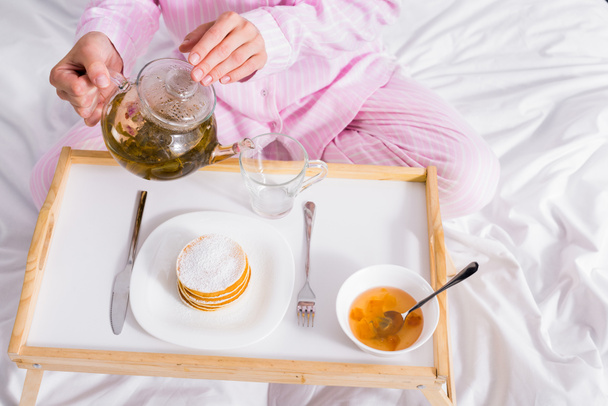 gedeeltelijke weergave van vrouw in roze pyjama groene thee in beker gieten terwijl het hebben van ontbijt in bed thuis - Foto, afbeelding
