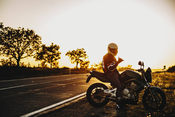 Increíble viajero en bicicleta caucásico sentado en su bicicleta cerca de la carretera y mirando a la cámara que muestra que está bien contra la puesta del sol
. - Foto, Imagen