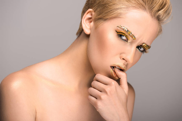 attraktive Frau mit goldenem Glanz im Gesicht beißt Daumen isoliert auf grau - Foto, Bild