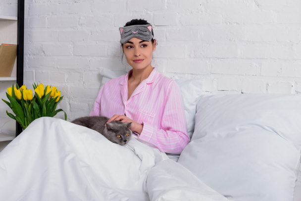 portrait of woman in pajamas and sleeping mask with britain shorthair cat resting in bed at home - Foto, afbeelding