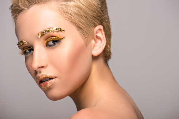 aantrekkelijke vrouw met gouden Glitters op gezicht kijken camera geïsoleerd op grijs - Foto, afbeelding