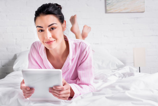 Porträt einer lächelnden Frau im Schlafanzug mit Tablet, während sie zu Hause im Bett liegt - Foto, Bild