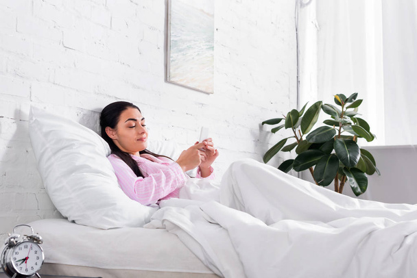 smiling woman in pajamas using smartphone while resting in bed at home - Foto, imagen