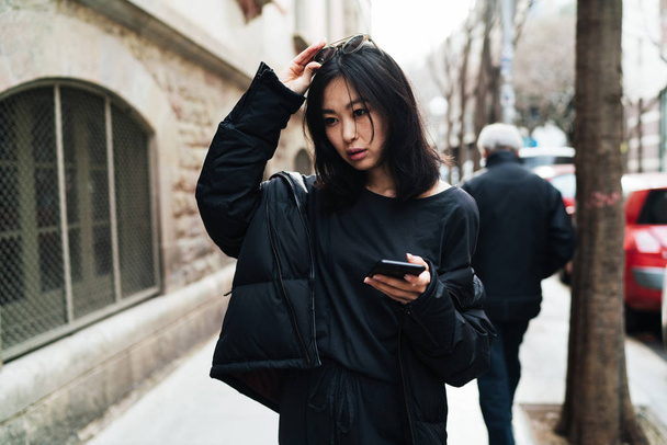 Beautiful asian woman in stylish outfit walking the street with a mobile phone in her hand. Hipster girl chatting online while going down the city - Photo, image