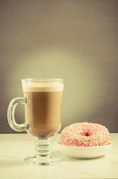 Coffee break: cappuccino glass with fresh sugary pink donut/Coffee break: cappuccino glass with fresh sugary pink donut on a gray background. Toned - Photo, Image