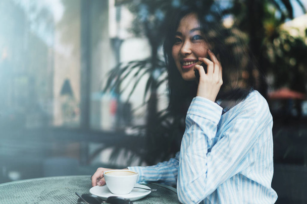 Joven hermosa mujer asiática hablando por un teléfono móvil mientras está sentado en una acogedora cafetería por la mañana
  - Foto, imagen