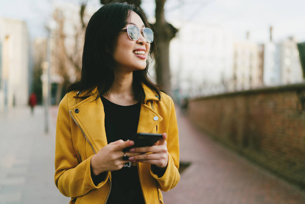 Half length portrait of a beautiful smiling asian female in a sunglasses with long dark hair wearing yellow leather jacket with mobile phone standing on blurred street background - Fotoğraf, Görsel