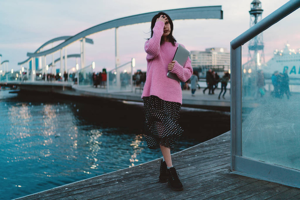 Full length portrait of stylish asian woman wearing pink sweater and chiffon dress adjusting her long dark hair while standing at the city berth in the evening with laptop - Foto, immagini
