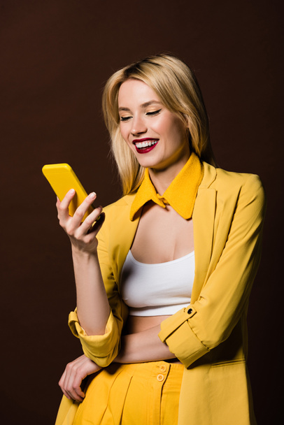 happy stylish blonde girl using yellow smartphone isolated on brown - Foto, afbeelding
