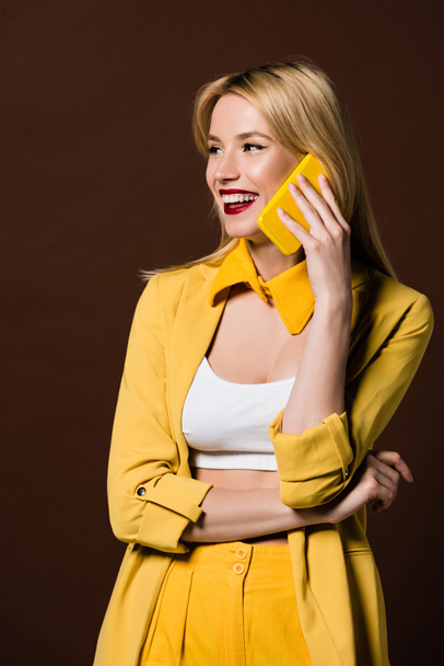 mulher loira bonita alegre falando por smartphone amarelo e olhando para longe isolado em marrom
 - Foto, Imagem