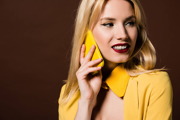 schöne stylische blonde Mädchen spricht mit gelben Smartphone und sieht weg isoliert auf braun - Foto, Bild