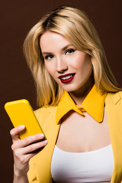 beautiful stylish blonde girl using yellow smartphone and smiling at camera isolated on brown - Fotografie, Obrázek