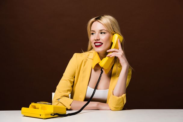 beautiful smiling blonde girl talking by vintage telephone and looking away on brown - Photo, Image