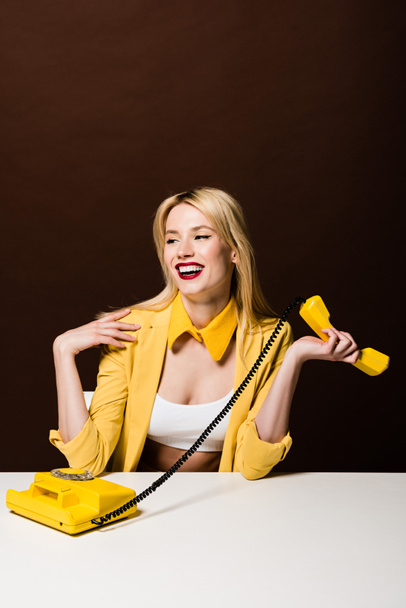 beautiful smiling blonde girl holding yellow handset and looking away on brown - Zdjęcie, obraz